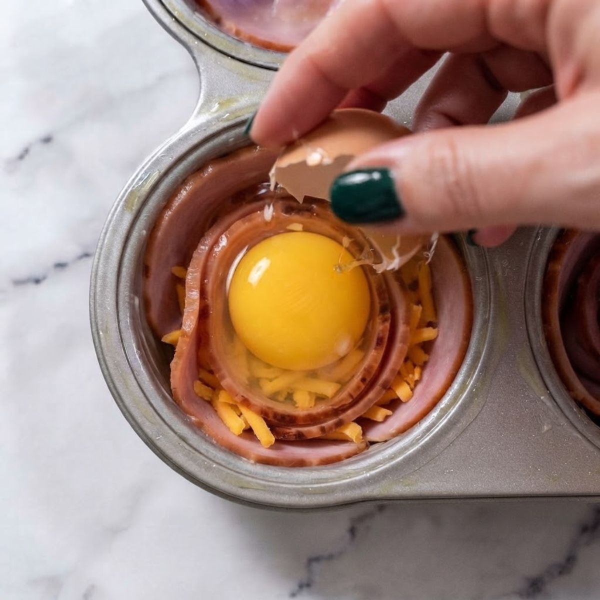 Extreme close-up shot of a woman with dark emerald green nails cracking an egg into a ham and cheese muffin cup on a marble counter.