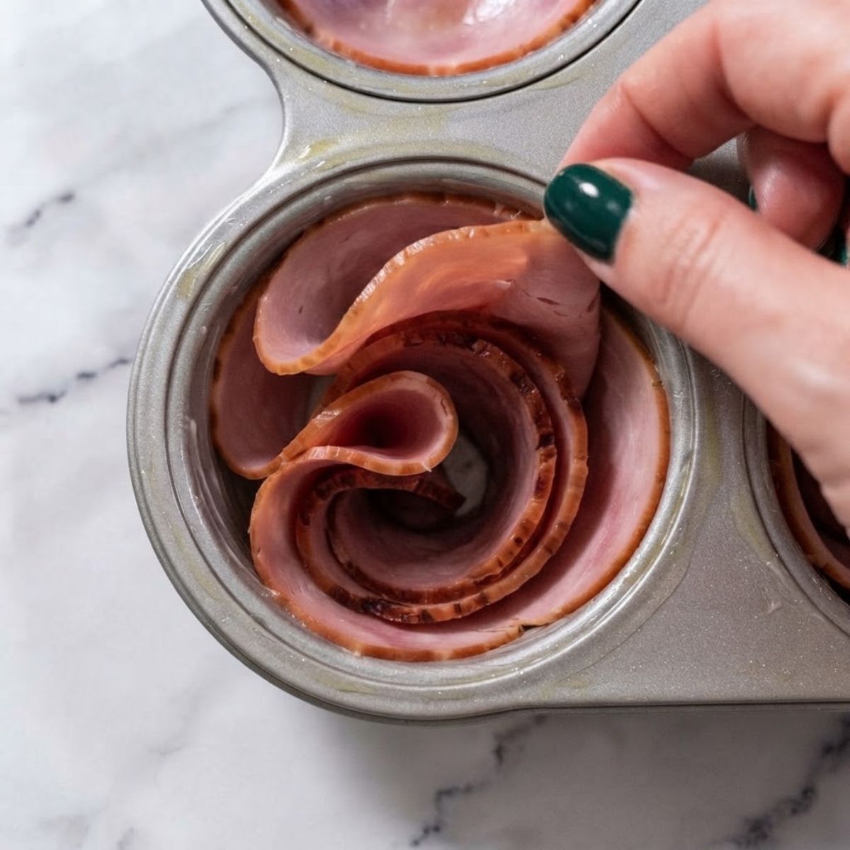 Extreme close-up shot of a woman with dark emerald green nails pressing a slice of deli ham into a greased muffin tin cup on a marble counter.