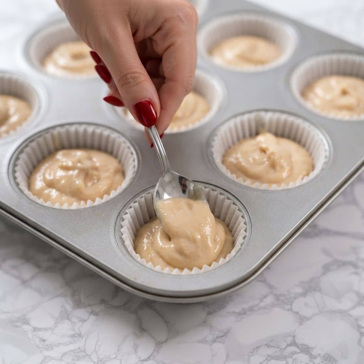 Extreme close-up shot of a woman with classic red nails spooning cupcake batter into paper liners in a muffin tin on a marble counter.