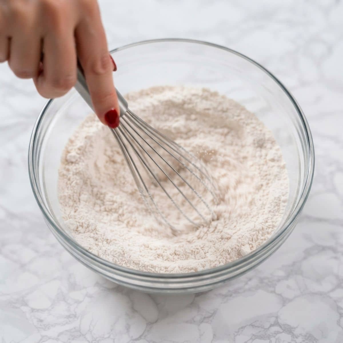 Extreme close-up shot of a woman with classic red nails whisking dry ingredients in a glass bowl on a marble counter.