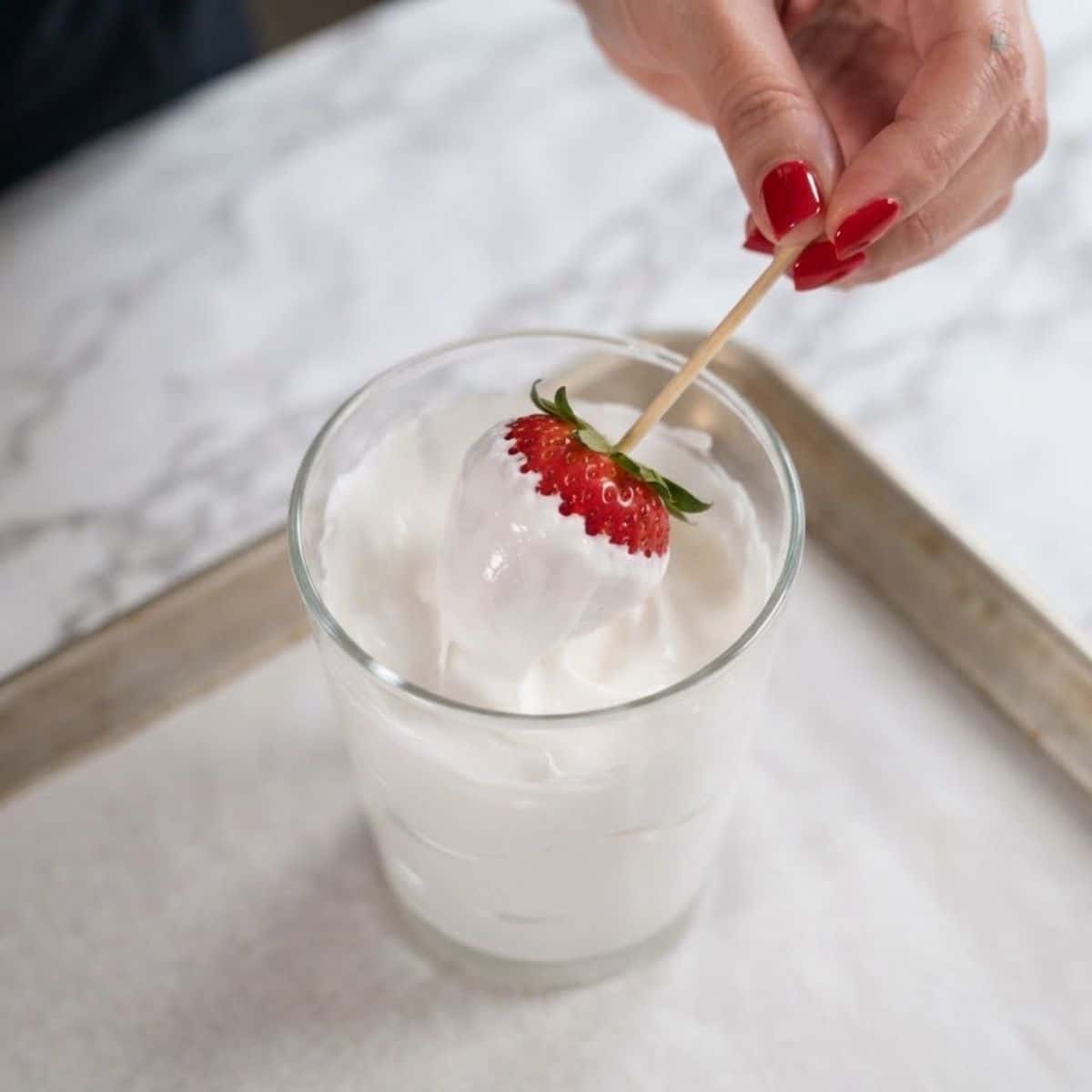Extreme close-up shot of a woman with classic red nails dipping a skewered strawberry into a tall glass bowl of marshmallow fluff on a marble counter.