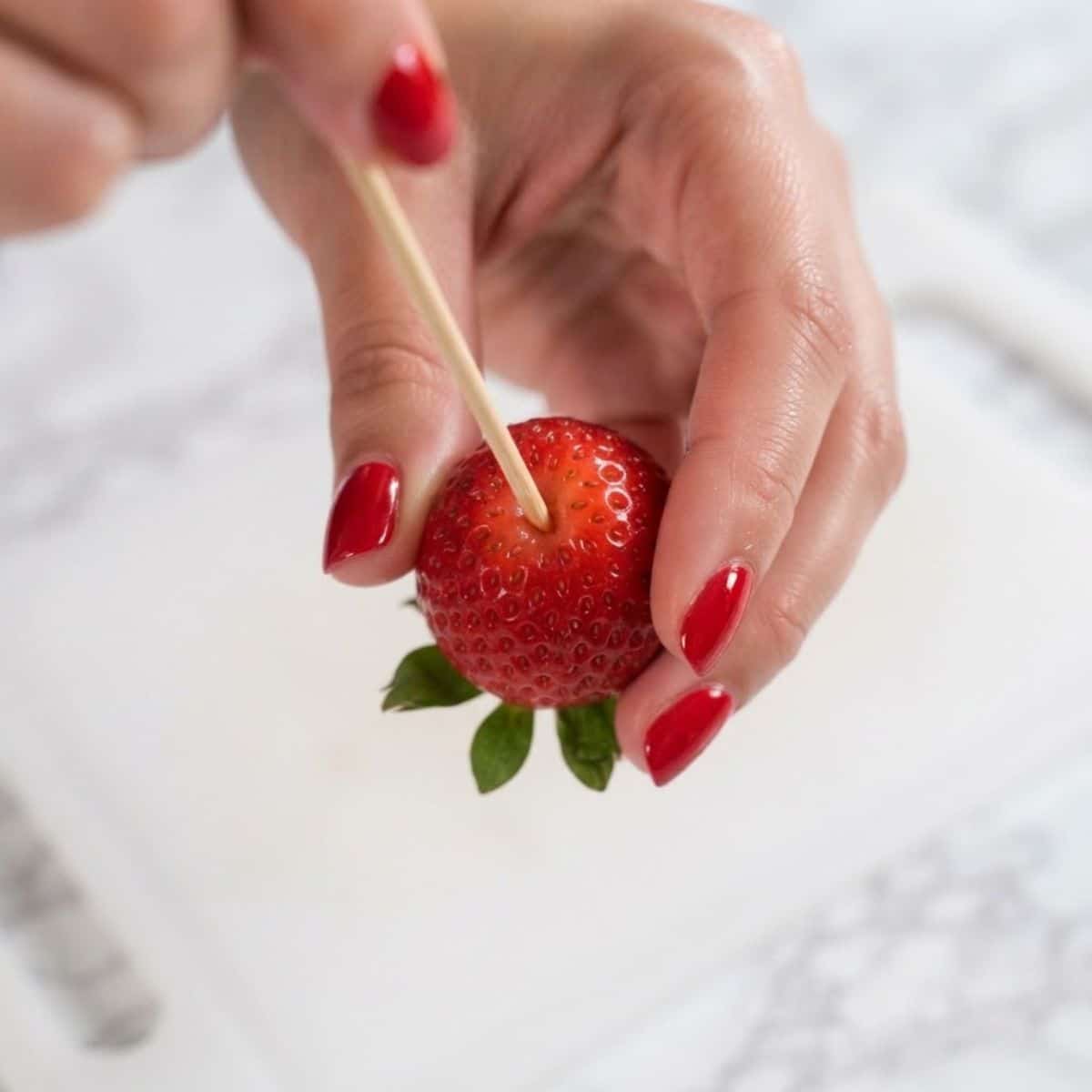 Extreme close-up shot of a woman with classic red nails inserting a wooden skewer into the top of a fresh strawberry on a cutting board.