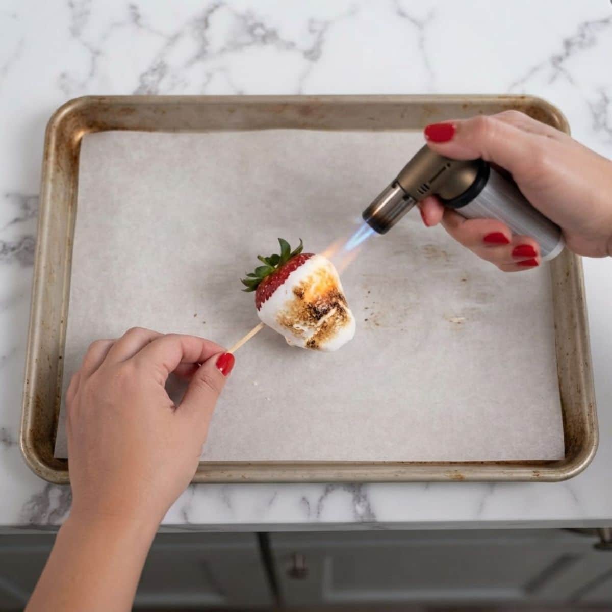 Extreme close-up shot of a woman with classic red nails using a kitchen torch to toast the marshmallow coating on a strawberry skewer over a marble counter.